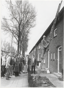 M 4254 Begin renovatie woningen Lokstraat Wethouder Grooten, met een dakpan in de hand op de ladder