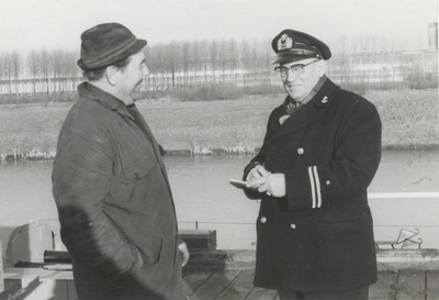 M 9086 Man in uniform schrijft lachend in bonnenboekje; andere man kijkt toe, staan beiden aan het water
