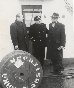 M 9091 Drie heren staand op een schip(?), een van hen draagt uniform, op de voorgrond een kabelhouder met opschrift ...