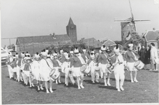 M 9251 Drumband in Lienden, spelend op een grasveld, kerk en molen op achtergrond