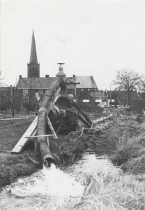 M 9260 Pijpleiding, uit de mond komt water dat in sloot wordt afgevoerd, op de achtergrond kerktoren en huizen