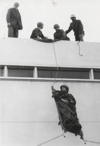 M 9287 Oefening hulpverleners of BB, vier mannen laten man op brancard naar beneden zakken uit gebouw
