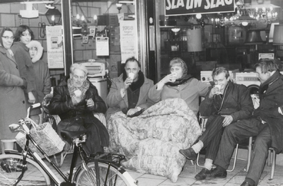 M 9331 Aantal mensen zittend en staand voor een winkel, twee hebben dekens om de voeten, voor een winkel met tekst 'Sla ...