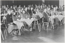 M 9865 Bijeenkomst, grote groep personen achter tafeltjes van formica, met kleedjes en koffiekopjes, luisterend