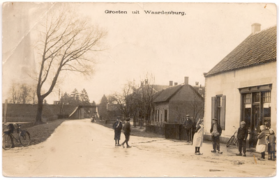 1099 Een prentbriefkaart van een straat in Waardenburg met rechts een kruidenierswinkel met daarvoor enkele kinderen. ...