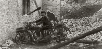 324 Jongetje op motorfiets tussen het puin van een afgebrand huis